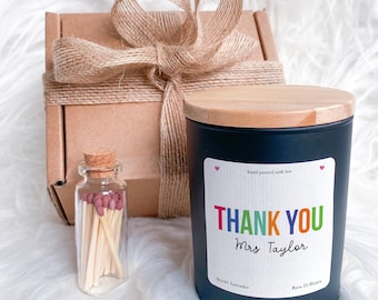 Op de afbeelding: Een zwarte kaars met een houten deksel en een label met de tekst "THANK YOU Mrs Taylor" in kleurrijke letters. Een klein glazen flesje met lucifers en een ingepakte geschenkdoos met een jute strik staan ook op de afbeelding. De geur van de kaars is lavendel.