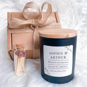 Puede incluir: Un juego de regalo con una vela negra con tapa de madera, una pequeña botella de cristal con cerillas y una caja envuelta. La vela tiene una etiqueta blanca con los nombres "Sophie & Arthur" y el texto "Will you be my Godparents?"