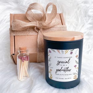 Op de afbeelding: Een cadeau set met een marineblauwe kaars met een houten deksel, een klein glazen flesje lucifers en een bruine geschenkdoos vastgebonden met touw. De kaars heeft een label met de tekst "It takes someone special to be a godmother".