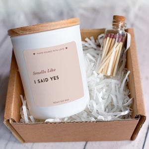 Könnte beinhalten: Eine weiße Kerze mit Holzdeckel und dem Aufdruck "Smells Like I Said Yes" befindet sich in einem Karton mit Papierstreifen. Eine kleine Glasflasche mit Streichhölzern befindet sich ebenfalls in der Box. Die Kerze ist mit "Hand Poured With Love" und "Vegan Soy Wax" beschriftet.