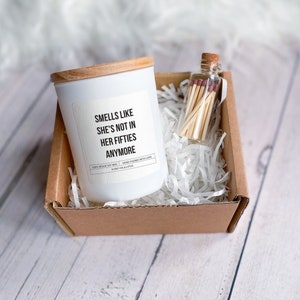 Könnte beinhalten: Eine weiße Kerze mit einem Holzdeckel in einem Karton mit weißem zerknülltem Papier. Die Kerze trägt den Text "Smells like she's not in her fifties anymore" auf dem Etikett. Die Schachtel enthält auch ein kleines Glas mit Streichhölzern.