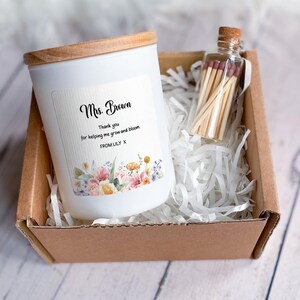 May include: A white candle with a wooden lid and a label that reads "Mrs. Brown" and a message. A small glass bottle of matches sits beside the candle in a cardboard box filled with white paper. The candle has a floral design.