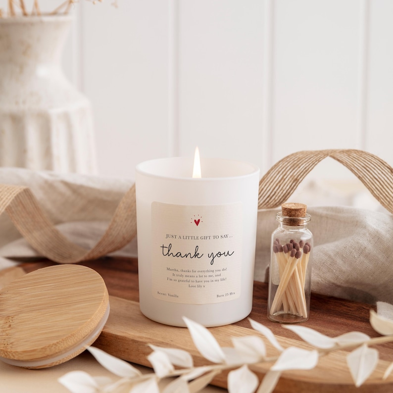 Peut inclure: Une bougie parfum&eacute;e blanche allum&eacute;e avec une &eacute;tiquette "merci" et une petite bouteille en verre d'allumettes. La bougie est pos&eacute;e sur un plateau en bois avec un couvercle en bois. C'est un cadeau.