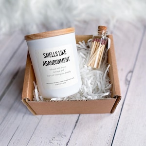 May include: A white candle in a glass jar with a wooden lid, featuring a label that reads "SMELLS LIKE ABANDONMENT" and "Infused with shock, betrayal and tears of missing you already". A small glass bottle of matches is included in the brown box.