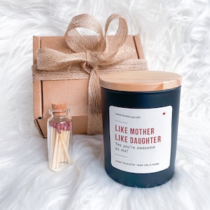 May include: A black candle with a wooden lid and a white label that reads "Like Mother Like Daughter Yes you're awesome as me!" The candle has a scent of fig and lotus and a burn time of 25 hours. The candle is packaged in a brown box with a burlap ribbon.