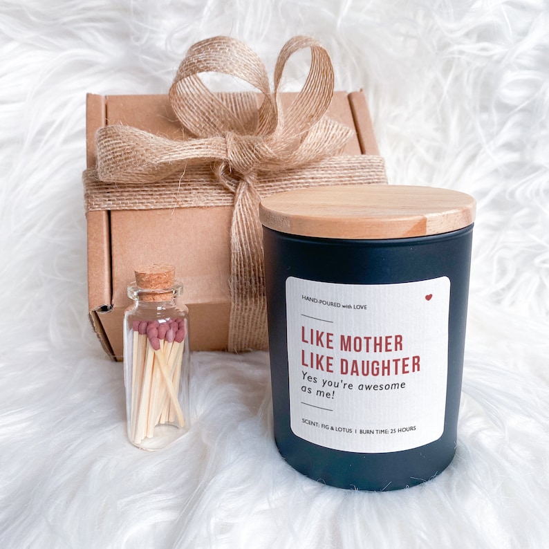 May include: A black candle with a wooden lid and a white label that reads "Like Mother Like Daughter Yes you're awesome as me!" The candle has a scent of fig and lotus and a burn time of 25 hours. The candle is packaged in a brown box with a burlap ribbon.