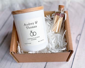 Puede incluir: Una vela blanca con una tapa de madera en una caja marrón con papel blanco triturado. La vela tiene una etiqueta que dice "Audrey & Thomas" con un icono de anillo y el texto "Felicidades 25.09.2025 Higo & Naranja Duración 35 Horas". Una pequeña botella de vidrio con un tapón de corcho llena de cerillas también está en la caja.