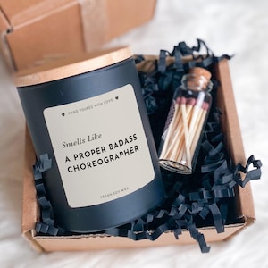 May include: A matte black candle jar with a wooden lid and a cream label reading Smells Like A PROPER BADASS CHOREOGRAPHER, accompanied by a small glass vial filled with wooden matches, nestled in a brown cardboard box with black paper filler.