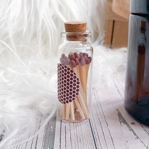 May include: A glass jar with a cork lid filled with wooden matches with brown tips. The jar has a red and white honeycomb pattern sticker on the side.