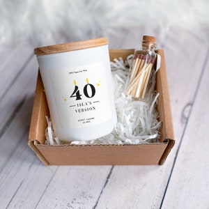 Könnte beinhalten: Eine weiße Kerze mit einem Holzdeckel in einer braunen Pappbox mit weißem zerknülltem Papier. Die Kerze hat ein Etikett mit der Aufschrift "40 Isla's Version" mit einer gelben Zahl 40 und zwei gelben Flammen. Die Kerze ist in Lavendelduft und brennt 15 Stunden lang. Ein kleines Glasgefäß mit einem Korkdeckel gefüllt mit Streichhölzern befindet sich ebenfalls in der Box.