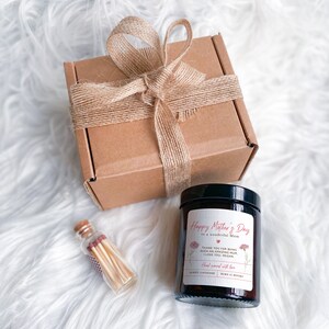 May include: A brown cardboard box with a burlap ribbon tied around it. The box is sitting on a white fluffy surface. Next to the box is a small glass jar with a brown lid. The jar contains a candle with a white label that says "Happy Mother's Day to a wonderful Mum". The label also includes a small red heart and the text "Thank you for being such an amazing Mum. I love you, Megan. Hand poured with love.  Berry Lavender. Burn 12 hours."
