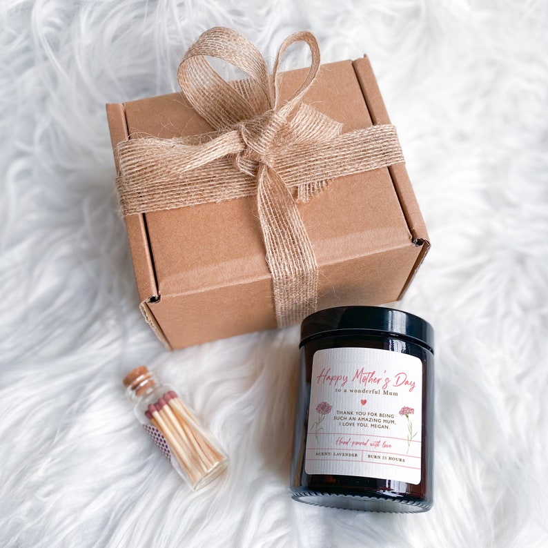 May include: A brown cardboard box with a burlap ribbon tied around it. The box is sitting on a white fluffy surface. Next to the box is a small glass jar with a brown lid. The jar contains a candle with a white label that says "Happy Mother's Day to a wonderful Mum". The label also includes a small red heart and the text "Thank you for being such an amazing Mum. I love you, Megan. Hand poured with love.  Berry Lavender. Burn 12 hours."