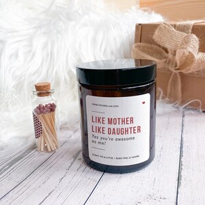 May include: A brown glass candle with a black lid. The label reads "Like Mother Like Daughter" and "Yes you're awesome as me!" The scent is fig and lotus and the burn time is 25 hours.