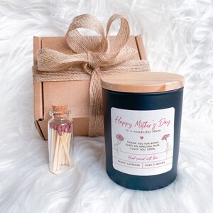 May include: A black candle with a wooden lid and a white label that reads "Happy Mother's Day to a wonderful Mum". The label also includes a pink heart and the text "Thank you for being such an amazing Mum. I love you. Emma." The candle is packaged in a brown box with a burlap ribbon. A small glass jar with matches is also included.