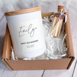 Könnte beinhalten: Eine weiße Kerze mit Holzdeckel, beschriftet mit "Emily" und "Happy 30th Birthday". Eine kleine Glasflasche mit Streichhölzern steht neben der Kerze in einer Pappschachtel mit weißem Papierfüllmaterial. Die Kerze duftet nach Lavendel und brennt 25 Stunden lang.