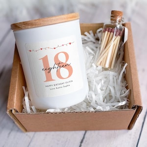 Puede incluir: Una vela blanca con tapa de madera y una etiqueta que dice "18 eighteen" en color melocotón. La vela está en una caja de cartón con papel triturado, junto a una pequeña botella de cristal con cerillas. La etiqueta también dice "Happy Birthday Olivia Love Auntie Sophie."