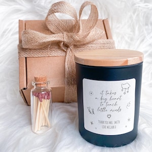 Pode incluir: Uma vela preta com tampa de madeira e um rótulo que diz "it takes a big heart to teach little minds". Também há uma pequena garrafa de vidro com fósforos de ponta rosa e uma caixa de presente amarrada com barbante.