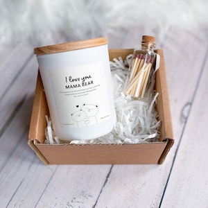Peut inclure: Une bougie blanche avec un couvercle en bois dans une boîte brune avec du papier blanc déchiqueté. La bougie porte une étiquette qui dit "I love you Mama Bear" avec un dessin de deux ours qui se font un câlin. Un petit bocal en verre avec des allumettes est également dans la boîte.