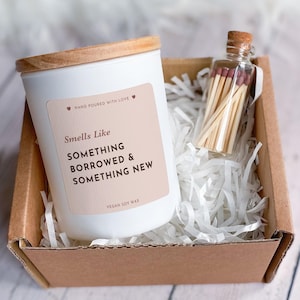 May include: A white candle with a wooden lid and a label that reads "Smells Like Something Borrowed & Something New" sits in a cardboard box with shredded paper. A small glass bottle filled with matches is also in the box. The candle is labeled "Vegan Soy Wax."