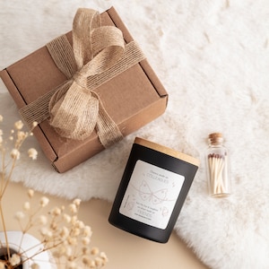 May include: A black candle with a wooden lid and a white label that says "Chance made us COLLEAGUES but the fun & laughter we share made us FRIENDS". The candle is on a white fluffy surface with a small glass bottle of matches and a brown box wrapped with twine.