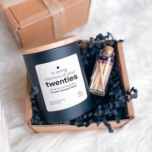 Könnte beinhalten: Eine schwarze Kerze mit Holzdeckel und einem Etikett mit der Aufschrift "In loving memory of your twenties". Die Kerze befindet sich in einer Pappschachtel mit schwarzem Papierfüllmaterial, zusammen mit einer kleinen Glasflasche mit Streichhölzern. Der Text "Gone but not forgotten forever young at heart" steht ebenfalls auf dem Etikett.