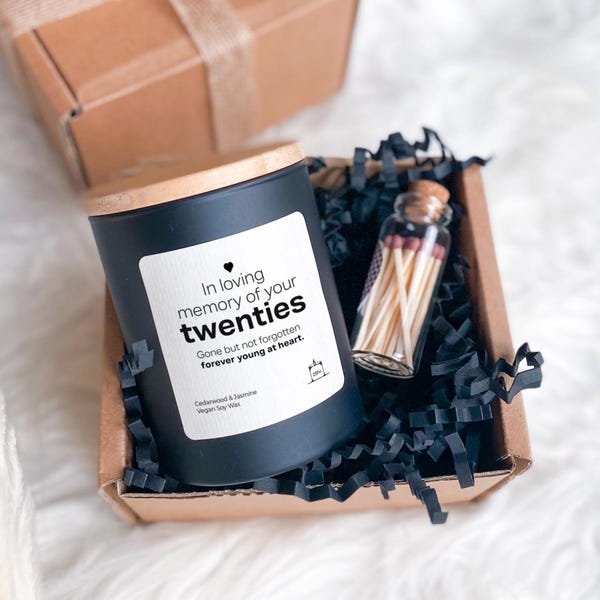Puede incluir: Una vela negra con tapa de madera y una etiqueta que dice "In loving memory of your twenties". La vela está en una caja de cartón con relleno de papel negro, junto a una pequeña botella de cristal de cerillas. El texto "Gone but not forgotten forever young at heart" también está en la etiqueta.