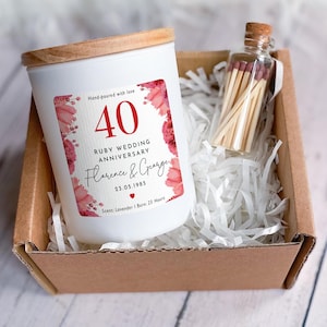 Puede incluir: Una vela blanca con tapa de madera y una etiqueta que dice "40 Aniversario de Bodas de Rubí Florence & George 23.05.1985". Una pequeña botella de vidrio de cerillas está junto a la vela en una caja de cartón.