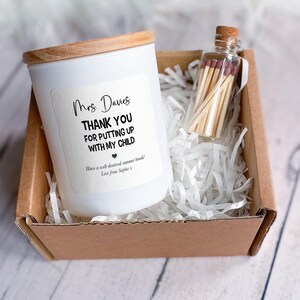 May include: A white candle with a wooden lid and a label that reads "Mrs. Davies THANK YOU FOR PUTTING UP WITH MY CHILD." A small glass bottle filled with matches sits next to the candle in a cardboard box filled with white paper shreds.