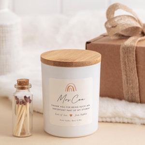 May include: A white candle with a wooden lid and a label that reads "Mrs. Cox" with a rainbow graphic. The text on the label says "THANK YOU FOR BEING SUCH AN IMPORTANT PART OF MY STORY! Lots of love from Sophie." A small glass bottle of matches is next to the candle.