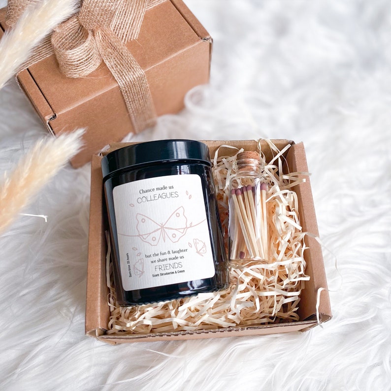May include: A gift box filled with brown shredded paper, containing a black candle with a white label that reads "Chance made us colleagues but the fun & laughter we share made us friends" and a small glass jar with matches. The candle has a butterfly design and the scent is "Sweet Strawberries & Cream".