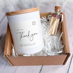 May include: A white candle with a wooden lid and a label that says "Thank you" in a gift box with matches. The candle is vanilla scented and has a 25-hour burn time. The gift box is filled with white paper shreds.