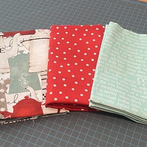 May include: Three folded fabric pieces with different patterns. One has a coffee theme with text "ÔTEL AFE", another is red with white polka dots, and the third is light green with coffee-related text.