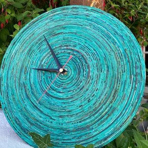 Puede incluir: Un reloj de pared redondo con un diseño en espiral turquesa y azul. Las manecillas del reloj son negras, con un segundero rojo. El reloj está hecho de papel enrollado y tiene una apariencia texturizada única. El reloj está al aire libre, rodeado de vegetación.
