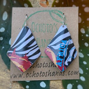 Puede incluir: Un par de pendientes en forma de diamante con un estampado de cebra y detalles coloridos. Los pendientes están sujetos a ganchos plateados y se exhiben en una tarjeta marrón con el texto "Ochoto's Hands" y una dirección web.