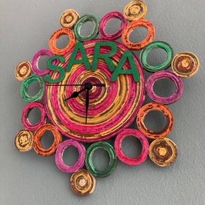 Puede incluir: Un reloj colorido hecho de papel enrollado con el nombre "SARA" en letras verdes en la parte delantera. El reloj tiene un diseño circular con círculos concéntricos de diferentes colores, incluyendo rosa, naranja, amarillo y verde.