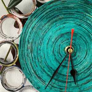 Può includere: Un quadrante di orologio blu turchese con lancette nere e rosse. Il quadrante dell'orologio è fatto di pezzi di carta arrotolati, creando un motivo circolare. L'orologio è circondato da un anello di carta arrotolata.