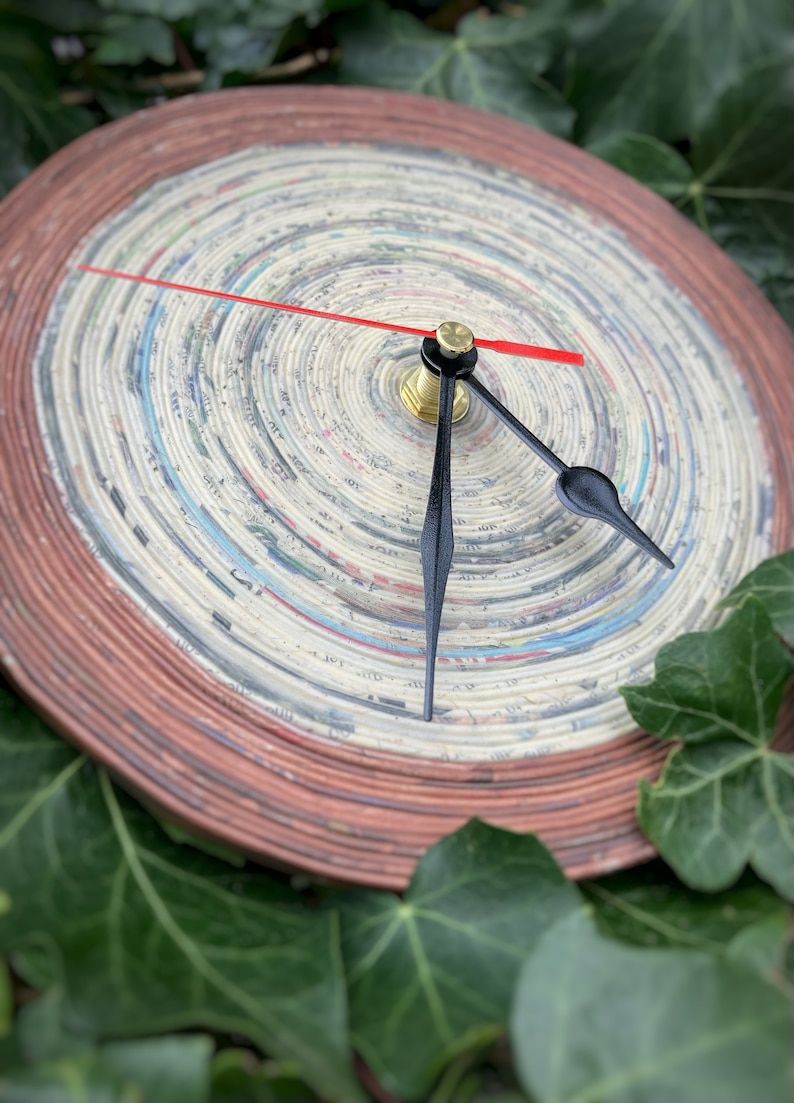 Op de afbeelding: Een ronde klok met een rode secondewijzer en zwarte uur- en minutenwijzers. De wijzerplaat van de klok is gemaakt van opgerolde krantenpagina's en heeft een bruine buitenring.