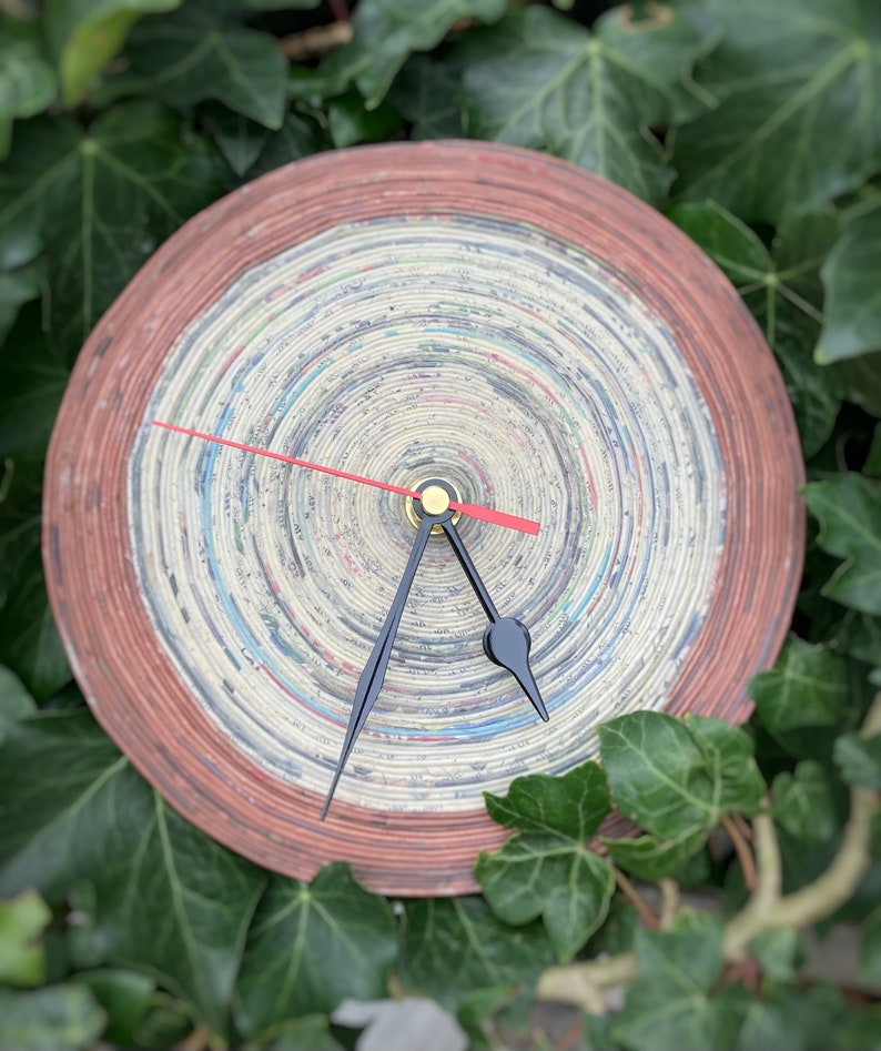 Op de afbeelding: Een ronde klok met een rode en bruine buitenrand. De wijzerplaat van de klok is gemaakt van gelaagd gerecycled papier, wat een uniek en getextureerd ontwerp cre&euml;ert. De wijzers van de klok zijn zwart met een gouden midden.