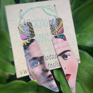 Puede incluir: Un par de pendientes únicos con un retrato de una mujer con lápiz labial rojo. Los pendientes tienen un diseño geométrico con un patrón colorido y están suspendidos de ganchos plateados. El fondo incluye el texto "choto hands".