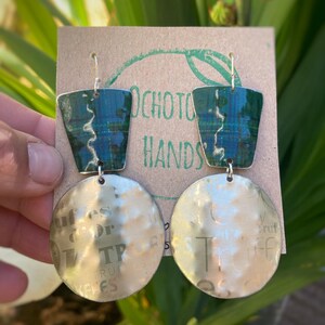 Puede incluir: Un par de pendientes con cuadrados de metal con estampado verde y azul y discos redondos plateados con texto. Los pendientes cuelgan de una tarjeta marrón con el texto "Ochotou Hands".