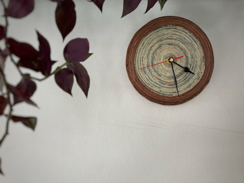 Op de afbeelding: Een ronde, bruine, papieren klok met een witte wijzerplaat en zwarte wijzers. De klok is aan een witte muur bevestigd.
