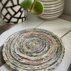 Puede incluir: Un posavasos redondo y colorido hecho de páginas de periódico enrolladas. El posavasos está hecho de muchas capas de papel, creando un patrón en espiral. El posavasos está sobre una superficie blanca.