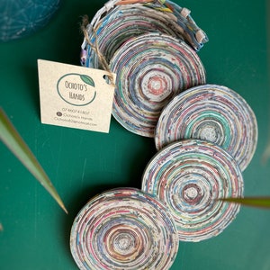 Könnte beinhalten: Set mit vier runden, bunten Untersetzern aus gerollten Recyclingpapier. Die Untersetzer haben ein konzentrisches Kreisdesign und sind perfekt zum Schutz von Oberflächen vor Hitze und Verschüttungen.
