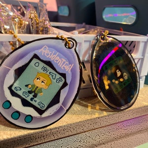 May include: A purple and blue plastic keychain shaped like a Tamagotchi with a digital screen displaying a cartoon character holding a gun. The screen also displays the text "Resident Evil".