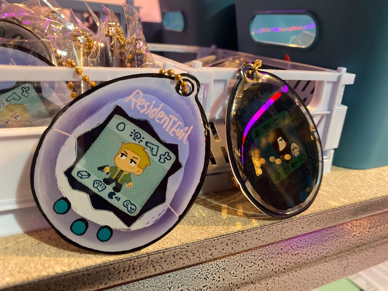 May include: A purple and blue plastic keychain shaped like a Tamagotchi with a digital screen displaying a cartoon character holding a gun. The screen also displays the text "Resident Evil".