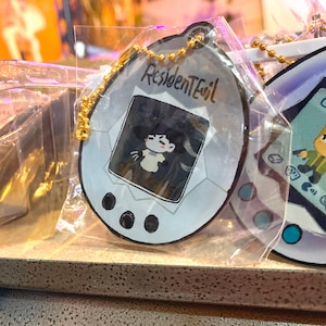 May include: A white and gray plastic keychain shaped like a digital pet with a black and white image of a character from the video game Resident Evil. The keychain has a gold chain attached to it.