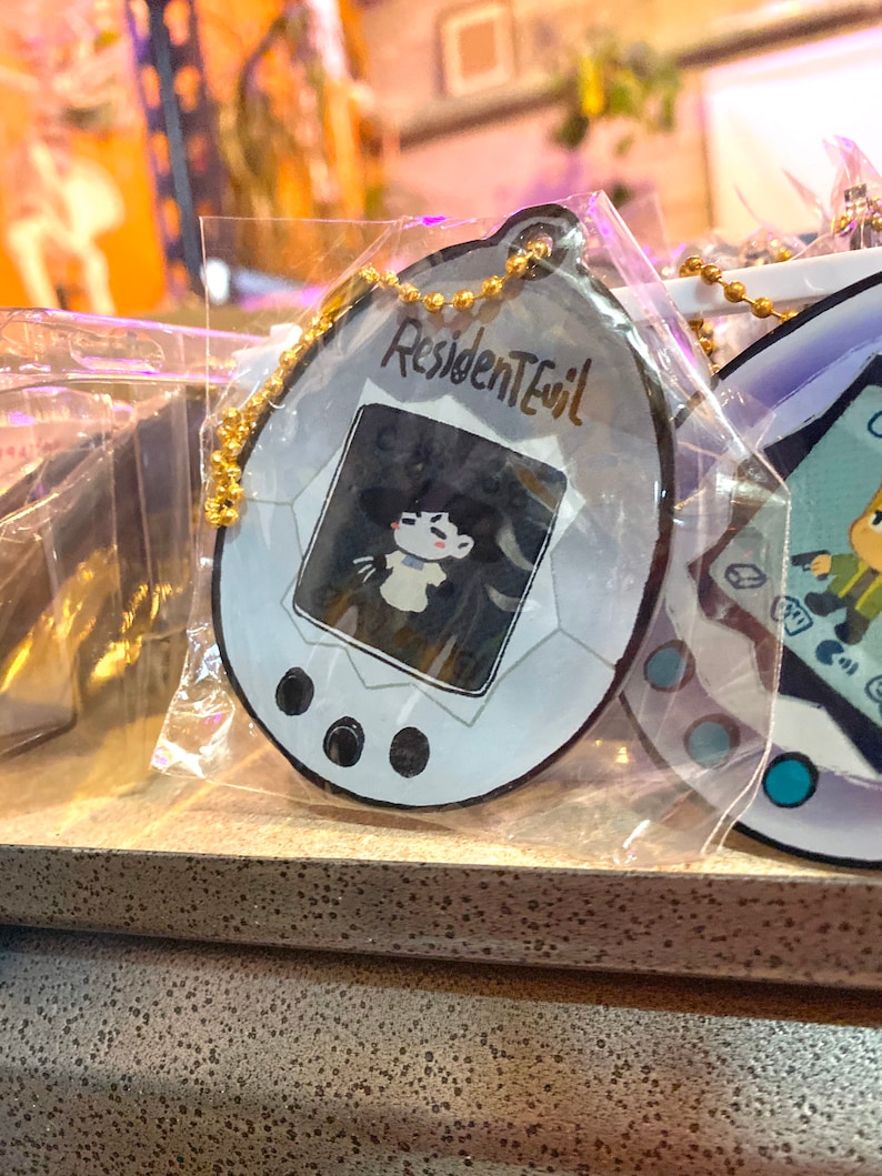 May include: A white and gray plastic keychain shaped like a digital pet with a black and white image of a character from the video game Resident Evil. The keychain has a gold chain attached to it.