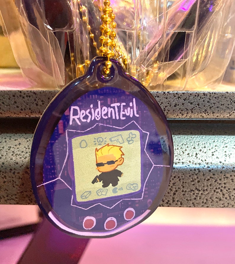May include: A purple and blue plastic keychain shaped like a digital pet with a cartoon image of a character from the video game Resident Evil. The keychain has the text "Resident Evil" on the top.
