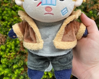 May include: A plush toy with blonde hair, a brown jacket, gray pants, and blue shoes. The toy has a white face with a red slash and blue bandages on its face.