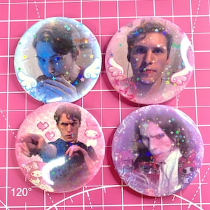 May include: Four round, holographic buttons with images of a person. Each button has a different image, with glitter and star accents. The buttons are on a pink grid background.